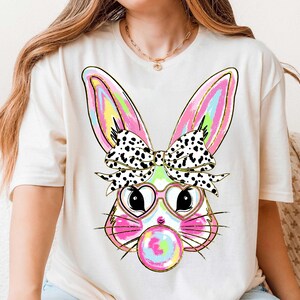 Puede incluir: Una camiseta blanca con un gr&aacute;fico colorido de un conejito que lleva gafas en forma de coraz&oacute;n y un lazo de lunares negros y blancos. El conejito est&aacute; soplando una burbuja de chicle.