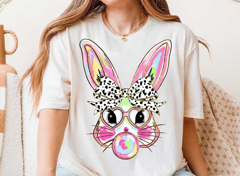 Puede incluir: Una camiseta blanca con un gr&aacute;fico colorido de un conejito que lleva gafas en forma de coraz&oacute;n y un lazo de lunares negros y blancos. El conejito est&aacute; soplando una burbuja de chicle.