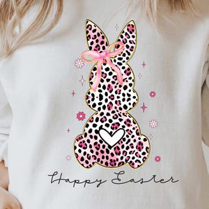 Puede incluir: Sudadera blanca con un conejo de estampado de leopardo rosa y negro que lleva un lazo rosa y el texto "Happy Easter".