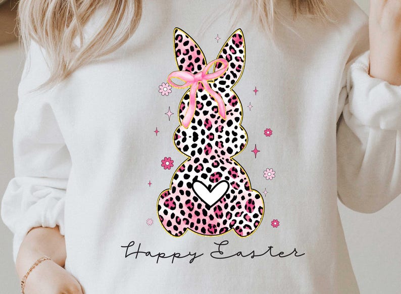 Puede incluir: Sudadera blanca con un conejo de estampado de leopardo rosa y negro que lleva un lazo rosa y el texto "Happy Easter".