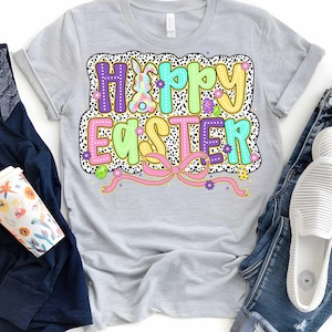 Puede incluir: Una camiseta gris claro con un gr&aacute;fico colorido de "Feliz Pascua" que presenta un conejito, flores y un lazo rosa. El gr&aacute;fico est&aacute; rodeado de un patr&oacute;n de lunares blanco y negro.