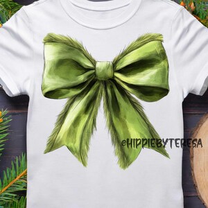 May include: White t-shirt featuring a large, detailed green bow graphic. The bow has a textured, realistic appearance. The text "HIPPIEBYTERESA" is printed on the lower right side of the shirt. The shirt is surrounded by Christmas decorations.