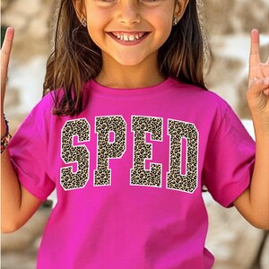 May include: A magenta t-shirt featuring the word "SPED" in large, bold letters. The letters have a leopard print pattern and white outlines. The shirt is worn by a young person.