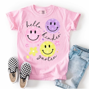 May include: Pink t-shirt with the words "hello kinder garter" and smiley face graphics. The shirt is paired with blue denim shorts and black and white checkered slip-on sneakers.