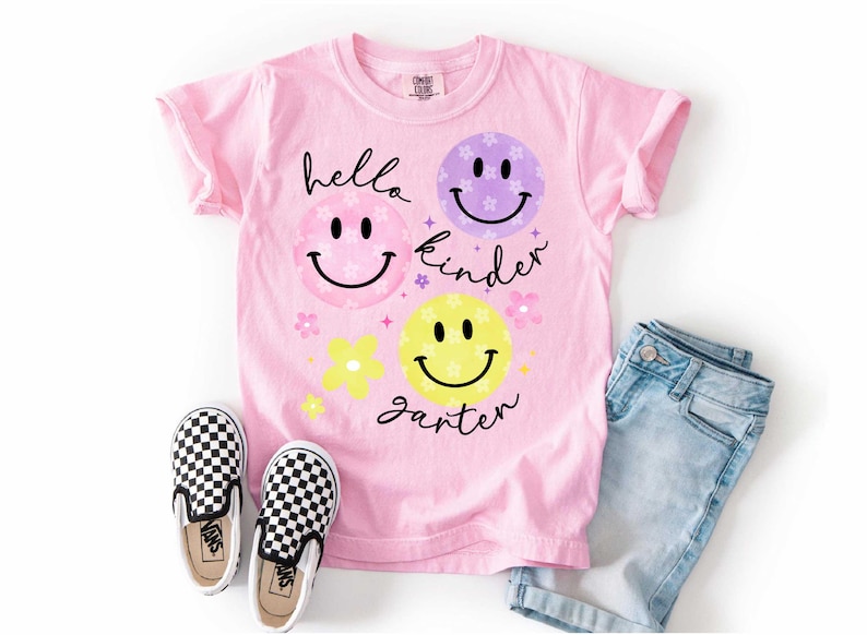 May include: Pink t-shirt with the words "hello kinder garter" and smiley face graphics. The shirt is paired with blue denim shorts and black and white checkered slip-on sneakers.