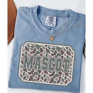May include: A light blue Comfort Colors t-shirt with a "MASCOT" graphic. The graphic features a camouflage pattern in shades of green, brown, and gray. A gold necklace with a pendant is draped over the shirt.