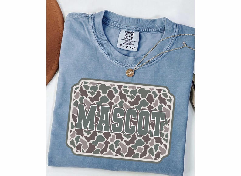 May include: A light blue Comfort Colors t-shirt with a "MASCOT" graphic. The graphic features a camouflage pattern in shades of green, brown, and gray. A gold necklace with a pendant is draped over the shirt.