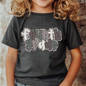 May include: Dark gray t-shirt with the words "Fourth Grade" in a playful, multi-colored font. The text has a camouflage-like pattern and is outlined in a darker shade. The shirt is short-sleeved and appears to be made of a soft material.