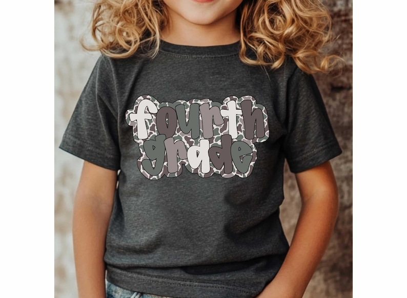 May include: Dark gray t-shirt with the words "Fourth Grade" in a playful, multi-colored font. The text has a camouflage-like pattern and is outlined in a darker shade. The shirt is short-sleeved and appears to be made of a soft material.