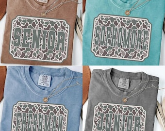 May include: Four Comfort Colors t-shirts in brown, teal, blue, and gray. Each shirt features a camo-patterned rectangle with a different grade level: "SENIOR", "SOPHOMORE", "FRESHMAN", and "JUNIOR". Each shirt has a gold necklace.