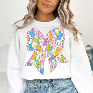 Puede incluir: Sudadera blanca con un dise&ntilde;o de lazo rosa. El lazo est&aacute; lleno de caramelos Peeps de colores.