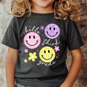 May include: Dark gray t-shirt featuring three smiley faces in pink, purple, and yellow, with the words "hello third grade" in white script. The design includes small flower accents.