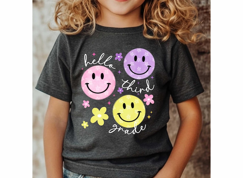 May include: Dark gray t-shirt featuring three smiley faces in pink, purple, and yellow, with the words "hello third grade" in white script. The design includes small flower accents.