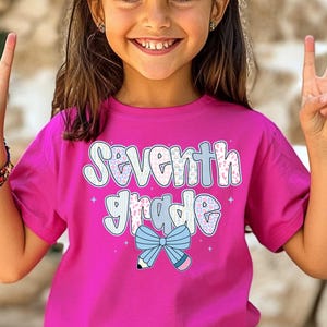 Pu&ograve; includere: Una maglietta rosa acceso con la scritta "Seventh Grade" in un font giocoso. Il design include un fiocco blu e una grafica a matita. Una scelta vivace per il ritorno a scuola o per l'uso quotidiano.