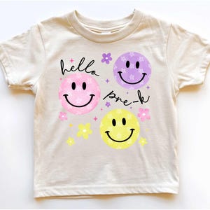 May include: Cream-colored t-shirt featuring a design with three smiley faces in pink, purple, and yellow, each with a smiling mouth and black eyes. The words "hello pre-k" are written in black script above and below the faces, with floral accents.