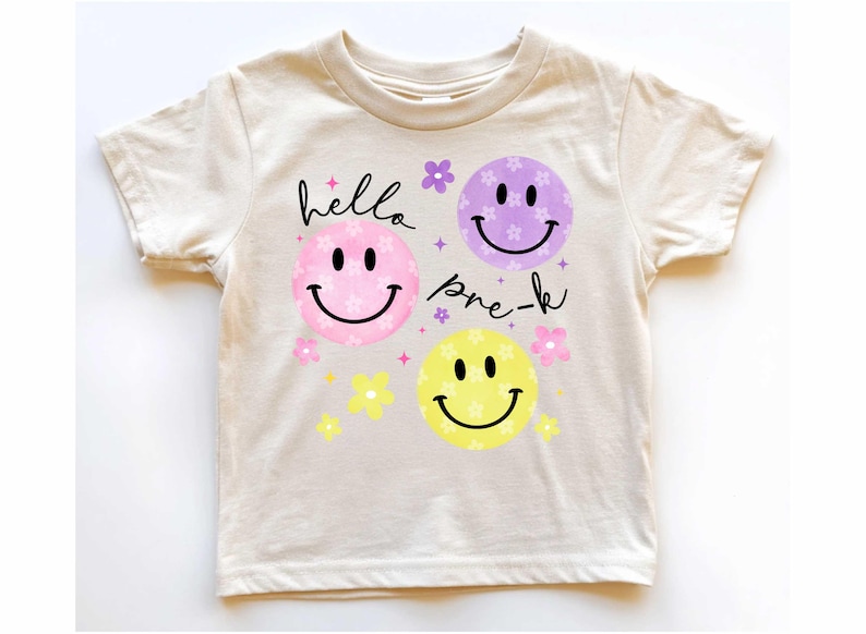 May include: Cream-colored t-shirt featuring a design with three smiley faces in pink, purple, and yellow, each with a smiling mouth and black eyes. The words "hello pre-k" are written in black script above and below the faces, with floral accents.