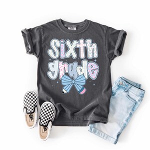 Pu&ograve; includere: Una maglietta grigio scuro con la scritta "Sixth Grade" in un carattere colorato e giocoso. La maglietta presenta un fiocco e una grafica a matita. L'immagine include anche pantaloncini di jeans e scarpe slip-on a scacchi, suggerendo un tema di ritorno a scuola.