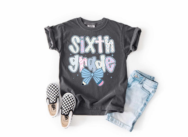 Pu&ograve; includere: Una maglietta grigio scuro con la scritta "Sixth Grade" in un carattere colorato e giocoso. La maglietta presenta un fiocco e una grafica a matita. L'immagine include anche pantaloncini di jeans e scarpe slip-on a scacchi, suggerendo un tema di ritorno a scuola.