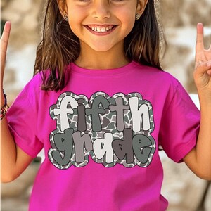 May include: A bright pink t-shirt with the words "fifth grade" in a playful, outlined font. The text has a camouflage pattern in shades of gray and white. The shirt is being worn by a young person.