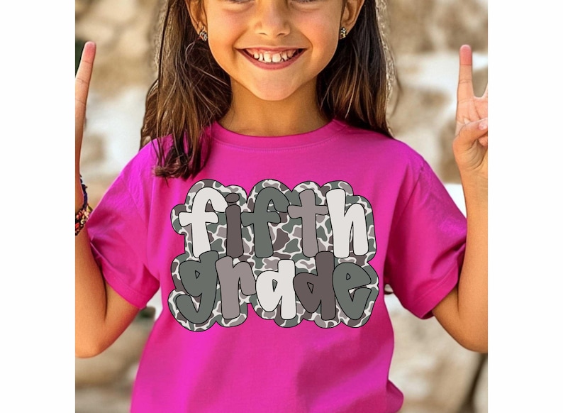 May include: A bright pink t-shirt with the words "fifth grade" in a playful, outlined font. The text has a camouflage pattern in shades of gray and white. The shirt is being worn by a young person.