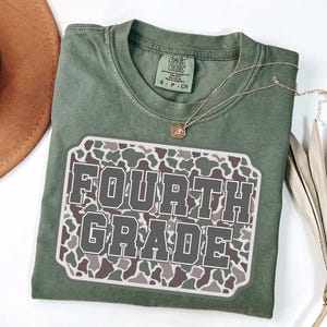 May include: A sage green t-shirt with a "FOURTH GRADE" graphic. The graphic is a rectangular patch with a camouflage pattern in shades of gray, brown, and white. A gold necklace with a pendant is on the shirt.