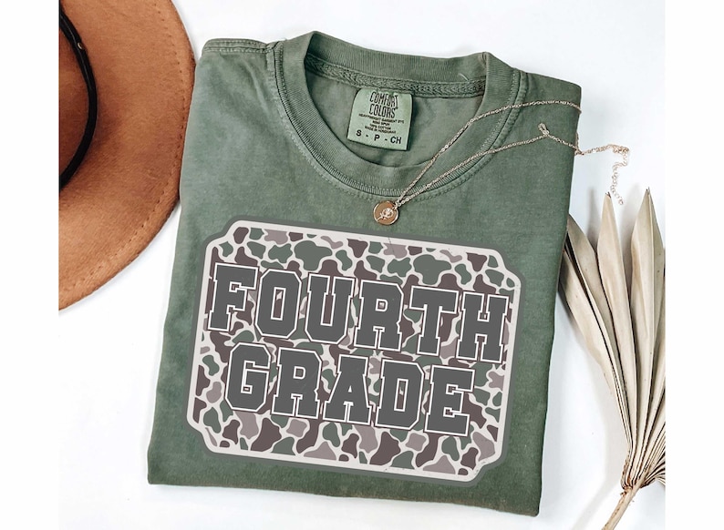 May include: A sage green t-shirt with a "FOURTH GRADE" graphic. The graphic is a rectangular patch with a camouflage pattern in shades of gray, brown, and white. A gold necklace with a pendant is on the shirt.