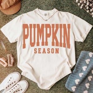 May include: Cream-colored t-shirt with the words "PUMPKIN SEASON" in orange block letters. The shirt is styled with white sneakers, a brown corduroy hat, and a silver necklace. A blue blanket with white hearts is also visible.