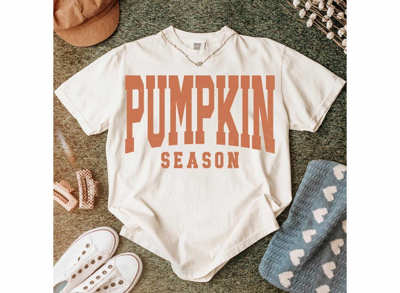 May include: Cream-colored t-shirt with the words "PUMPKIN SEASON" in orange block letters. The shirt is styled with white sneakers, a brown corduroy hat, and a silver necklace. A blue blanket with white hearts is also visible.