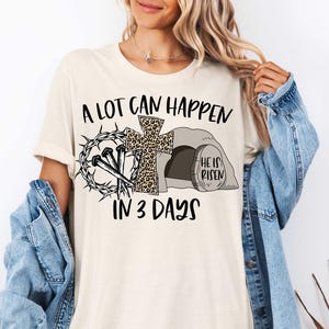 Puede incluir: Una camiseta blanca con un dise&ntilde;o gr&aacute;fico negro que dice "A lot can happen in 3 days". El gr&aacute;fico incluye una cruz con un dise&ntilde;o de estampado de leopardo, clavos, una corona de espinas y una tumba con las palabras "He is risen".