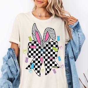 Puede incluir: Una camiseta blanca con un lazo a cuadros blanco y negro con orejas de conejo rosas. El lazo est&aacute; rodeado de formas de conejitos de Pascua de colores.