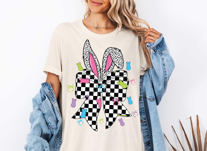 Puede incluir: Una camiseta blanca con un lazo a cuadros blanco y negro con orejas de conejo rosas. El lazo est&aacute; rodeado de formas de conejitos de Pascua de colores.