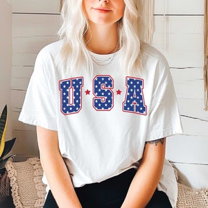 May include: White t-shirt with a red, white, and blue "USA" graphic. The letters are outlined in red and filled with blue and white stars.
