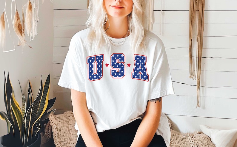 May include: White t-shirt with a red, white, and blue "USA" graphic. The letters are outlined in red and filled with blue and white stars.