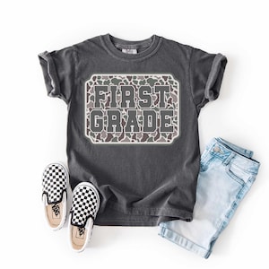 May include: A charcoal gray t-shirt with the words "FIRST GRADE" in a camo design. The shirt is paired with light blue denim shorts and black and white checkered slip-on sneakers.