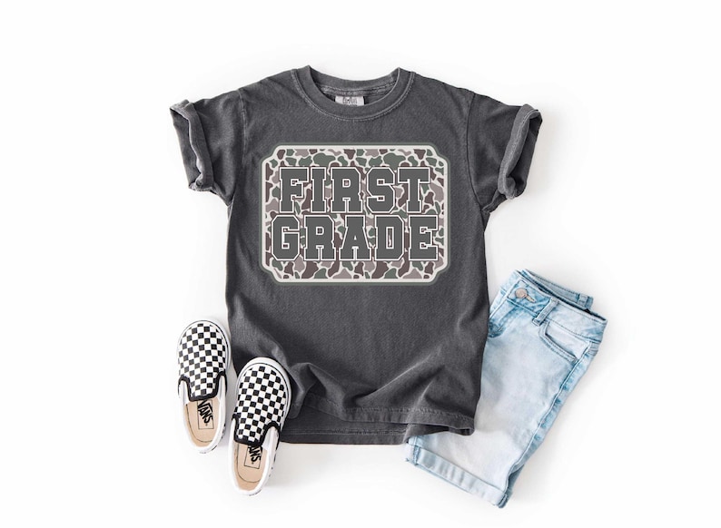 May include: A charcoal gray t-shirt with the words "FIRST GRADE" in a camo design. The shirt is paired with light blue denim shorts and black and white checkered slip-on sneakers.