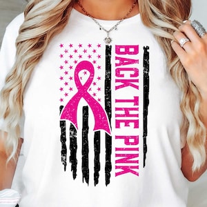 Puede incluir: Camiseta blanca con una cinta rosa y un diseño de bandera estadounidense. El texto "Back the Pink" está impreso verticalmente en la bandera.