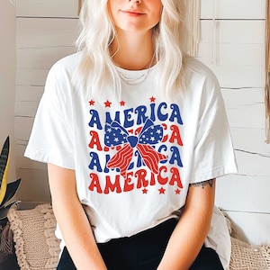May include: White t-shirt with a red, white, and blue bow and the word "AMERICA" repeated four times in a retro font.