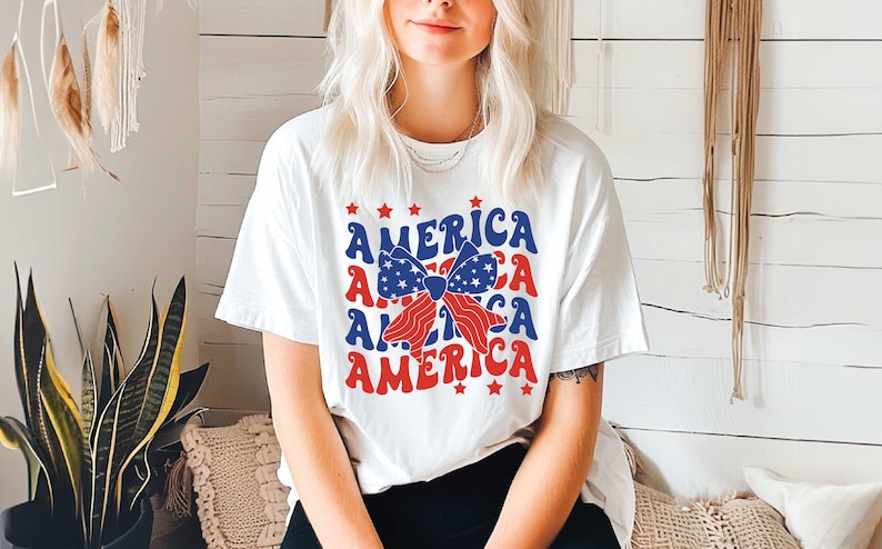 May include: White t-shirt with a red, white, and blue bow and the word "AMERICA" repeated four times in a retro font.
