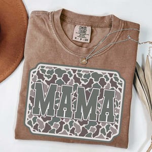 May include: A brown Comfort Colors t-shirt with a large graphic that says "MAMA" in gray letters. The graphic has a camouflage pattern in shades of green, gray, and brown. A silver necklace and a brown hat are also visible.