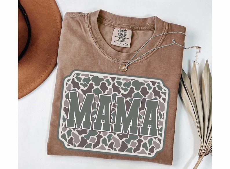 May include: A brown Comfort Colors t-shirt with a large graphic that says "MAMA" in gray letters. The graphic has a camouflage pattern in shades of green, gray, and brown. A silver necklace and a brown hat are also visible.