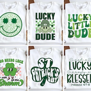 May include: Six white t-shirts with green shamrock and St. Patrick's Day themed designs. The designs include a smiling shamrock, the text "Lucky Dude", "Lucky Little Dude", "Who Needs Luck with All This Charm", "St. Patrick's Day", and "Not Lucky Simply Blessed Romans 4:7-8".