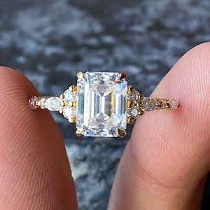 Könnte beinhalten: Ein goldener Verlobungsring mit einem Smaragdschliff-Diamanten als Mittelstück und kleineren Diamanten auf der Ringschiene.