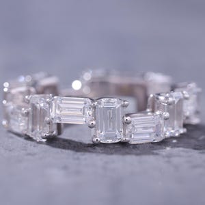 Puede incluir: Un anillo plateado con múltiples piedras preciosas rectangulares y transparentes. El anillo está sobre una superficie gris. Las piedras son de talla esmeralda.