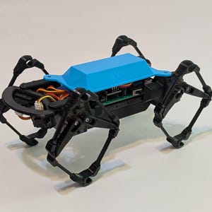 以下が含まれることがあります： 関節のある脚を持つ、黒と青のロボットスパイダー。ロボットは青いトップカバーと、ワイヤーや回路基板を含む、目に見える内部コンポーネントを備えています。脚はクモの動きを模倣するように設計されています。