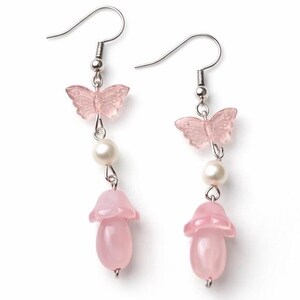 Könnte beinhalten: Ohrringe zum Aufhängen mit einem rosa Schmetterling, einer weißen Perle und einem rosa Pilzanhänger. Die Ohrringe haben silberfarbene Haken und Verbindungsbeschläge. Die Schmetterlings- und Pilzanhänger sind durchscheinend.