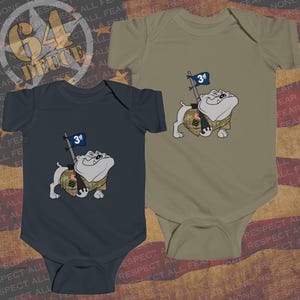 May include: Two baby onesies, one navy blue and one olive green. Each features a cartoon bulldog wearing a camouflage vest and holding a flag with "3d" on it. The background includes text and a star graphic.