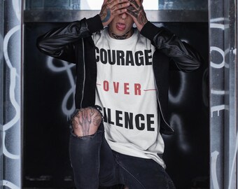 May include: White t-shirt with the words "COURAGE OVER SILENCE" in black and red. The person is wearing a black jacket and black jeans. The background has graffiti.