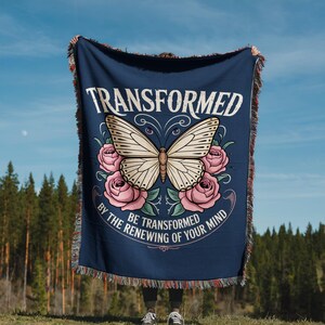 May include: A navy blue throw blanket with a butterfly and rose design. The blanket features the words "TRANSFORMED" and "BE TRANSFORMED BY THE RENEWING OF YOUR MIND" in white script. The edges of the blanket have a fringed trim.