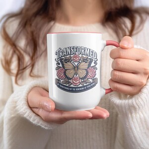 May include: White ceramic mug with a pink interior and handle. The mug features a butterfly design with the word "TRANSFORMED" above it, surrounded by roses. The phrase "BE TRANSFORMED BY THE RENEWING OF YOUR MIND" is below the butterfly.