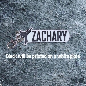 May include: A white keychain with the name "ZACHARY" in black block letters. A black silhouette of a person performing a martial arts kick is on the left. The text "Black will be printed on a white plate" is below.